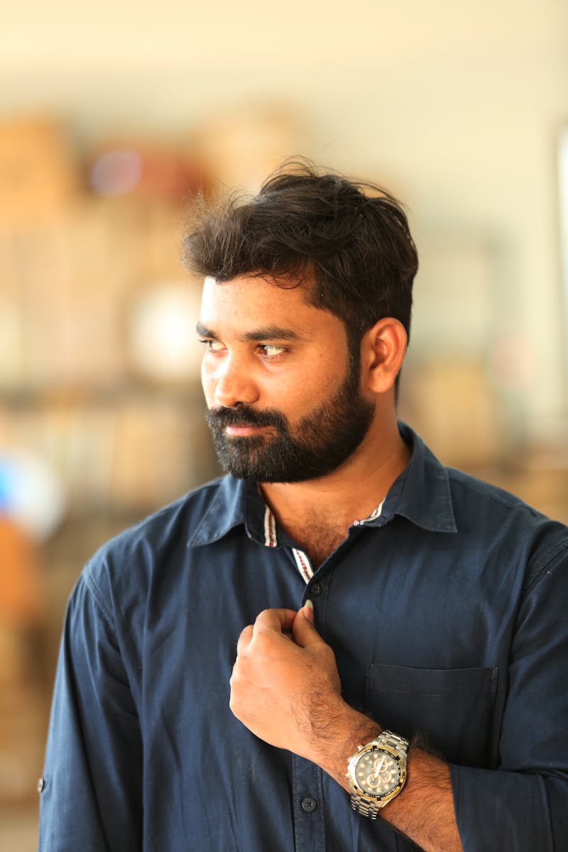 A thoughtful man with a beard and blue shirt posing indoors, displaying a stylish watch.