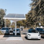 A white electric car charging under a solar panel in an urban setting, showcasing renewable energy use.